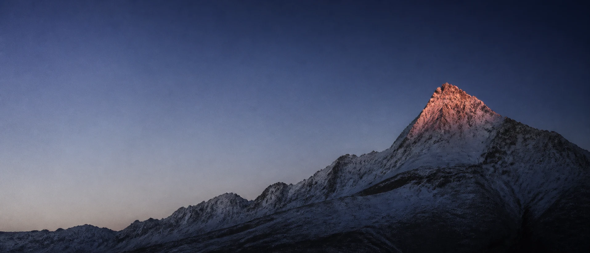 A high summit holding the last rose-gold light while the valley falls into blue shadow